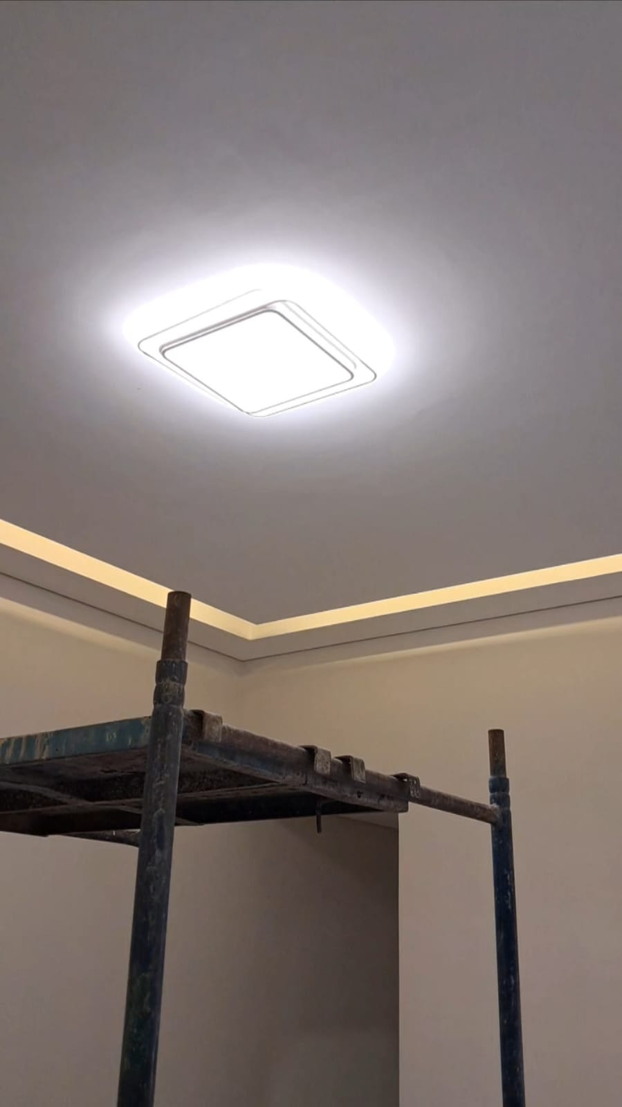 foto de um ambiente interno com luminária quadrada de teto acesa, sancas de gesso com fita de LED e andaime me